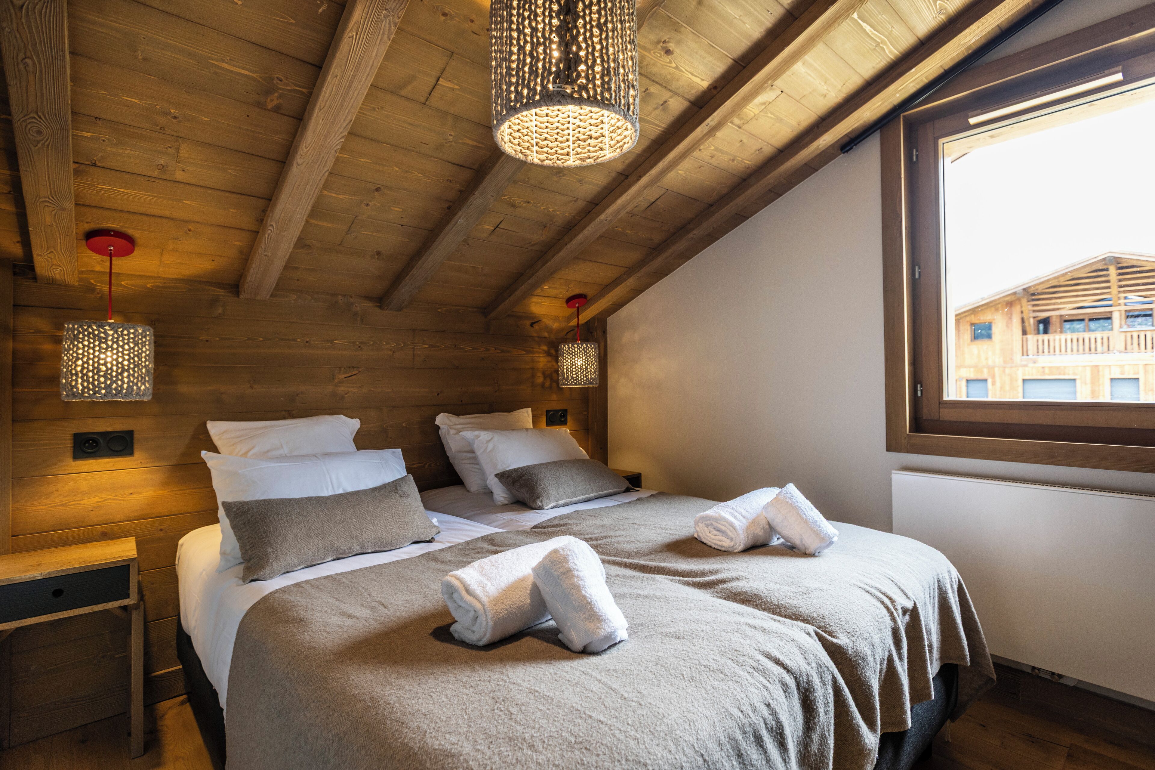 Een knus slaapkamer met houten balken, twee bedden met handdoeken, en een raam met uitzicht op een gebouw.