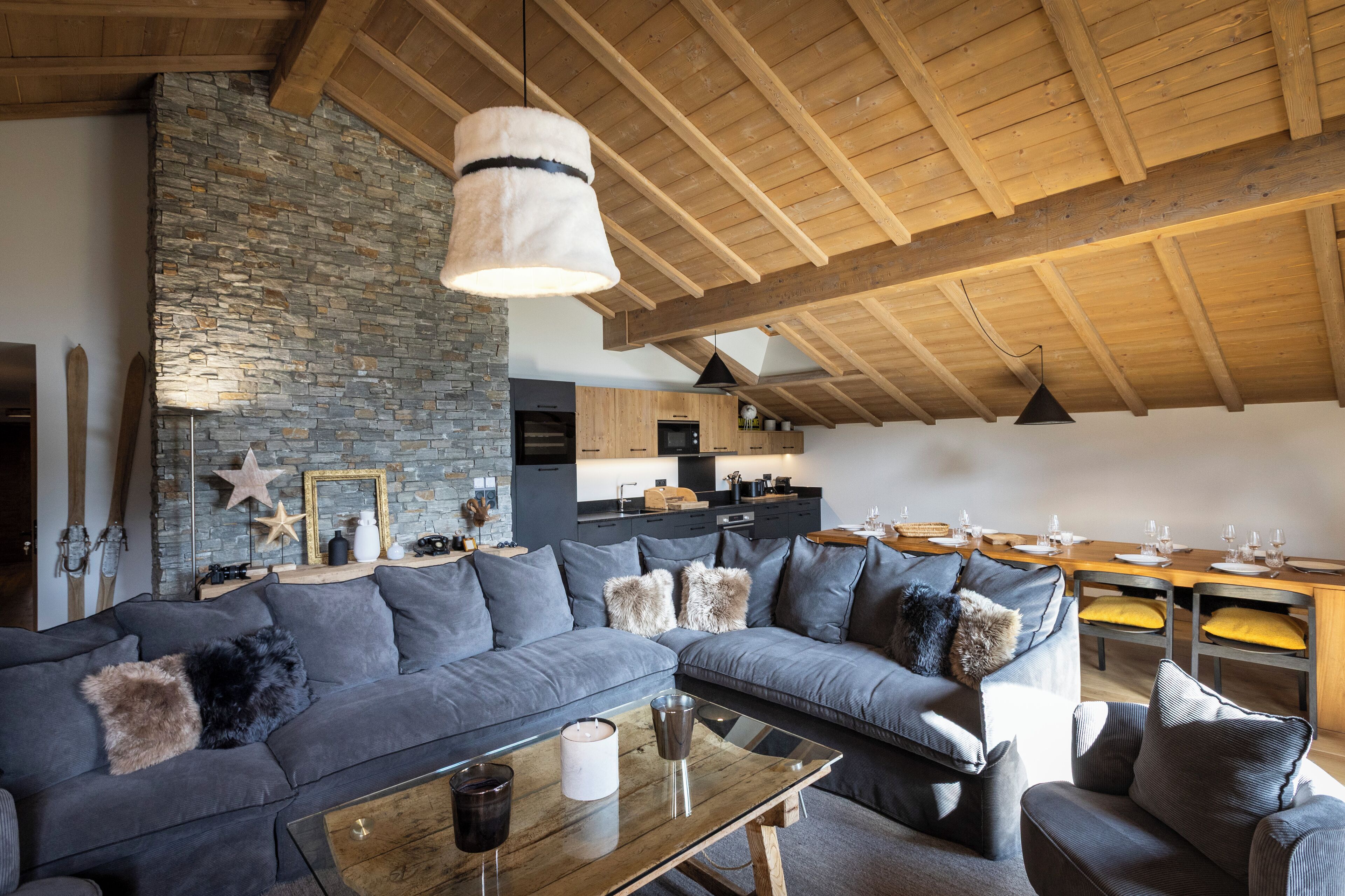 A modern living room with a stone wall, a wooden ceiling, and a dining area with yellow chairs.
