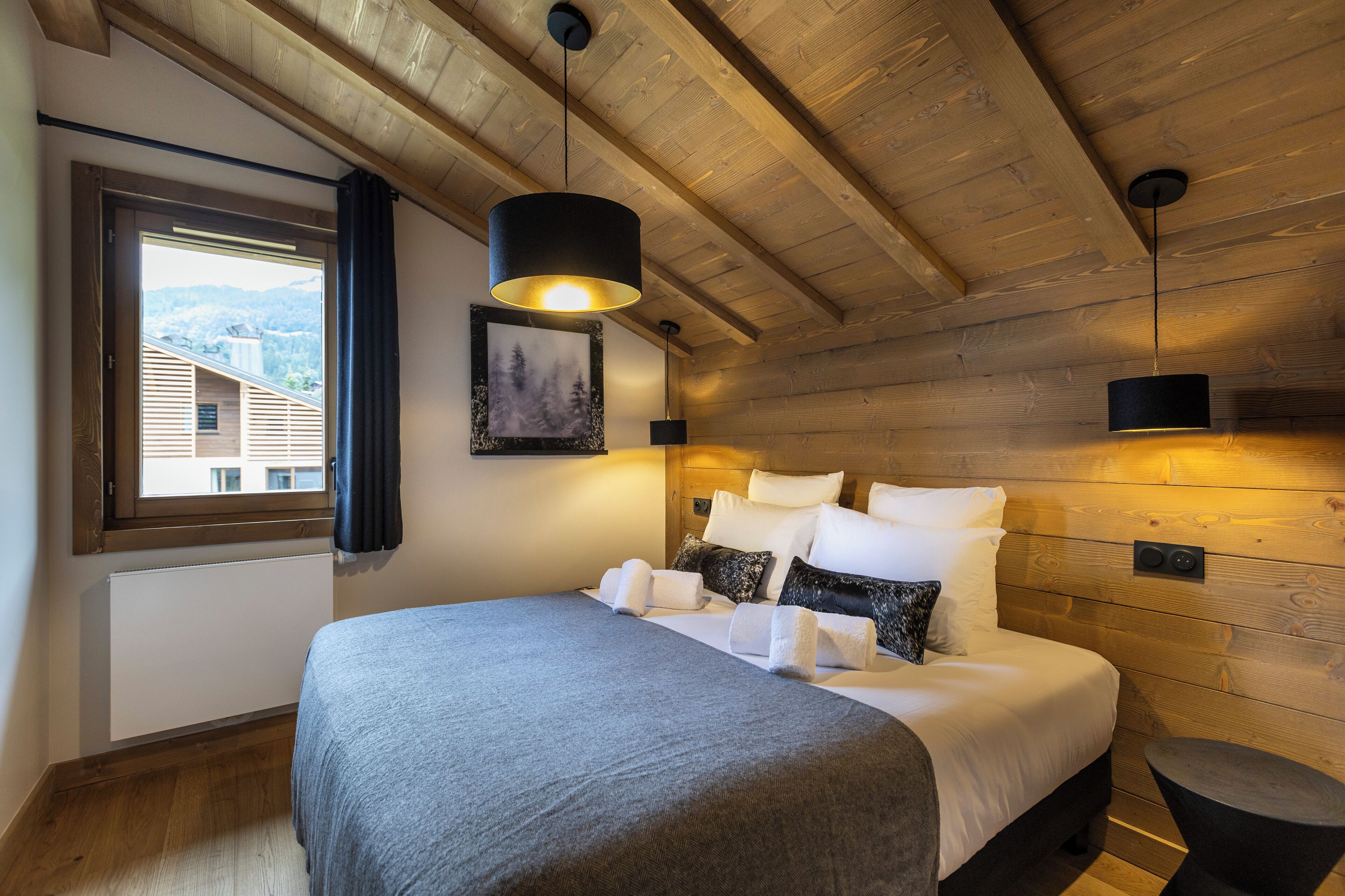 A modern bedroom with a wooden ceiling, a large bed with white linens, a black hanging light fixture, and a window with a view of buildings.