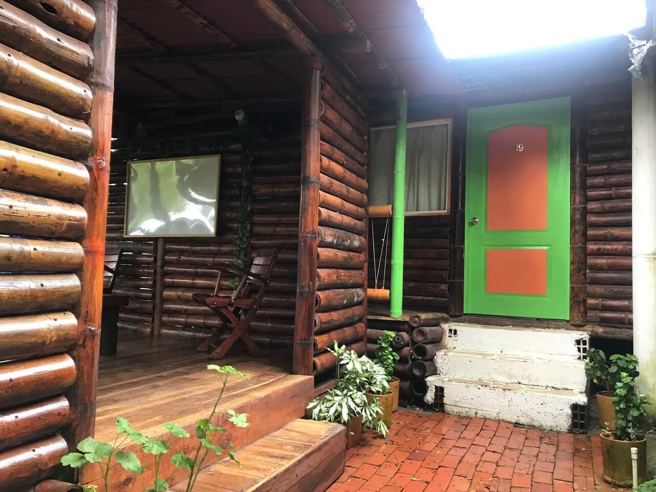 A log cabin with a green door, wooden steps, and a porch area with chairs and potted plants.