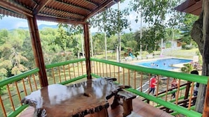 A wooden table with a natural edge design, a green metal railing, and a view of a swimming pool and greenery in the background. - Ecocabañas Pereira (Pereira)