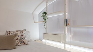 A bedroom with a bed, a bedside table, a hanging plant, and a window with blinds.