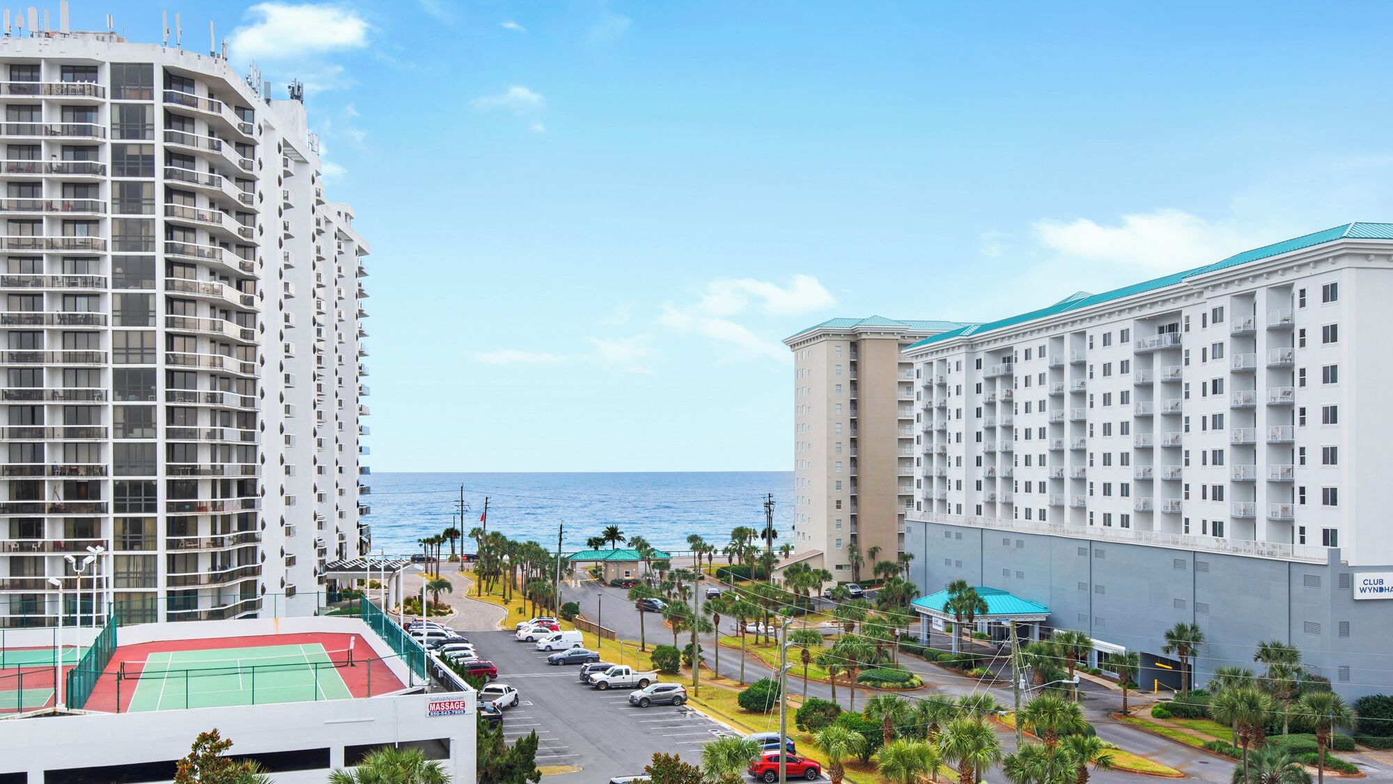 A high-rise hotel with multiple balconies, a tennis court, and a view of the ocean.