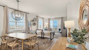 Uma sala de estar ampla com uma mesa de jantar de madeira, cadeiras e um espelho grande. Há uma varanda com vista para o mar.