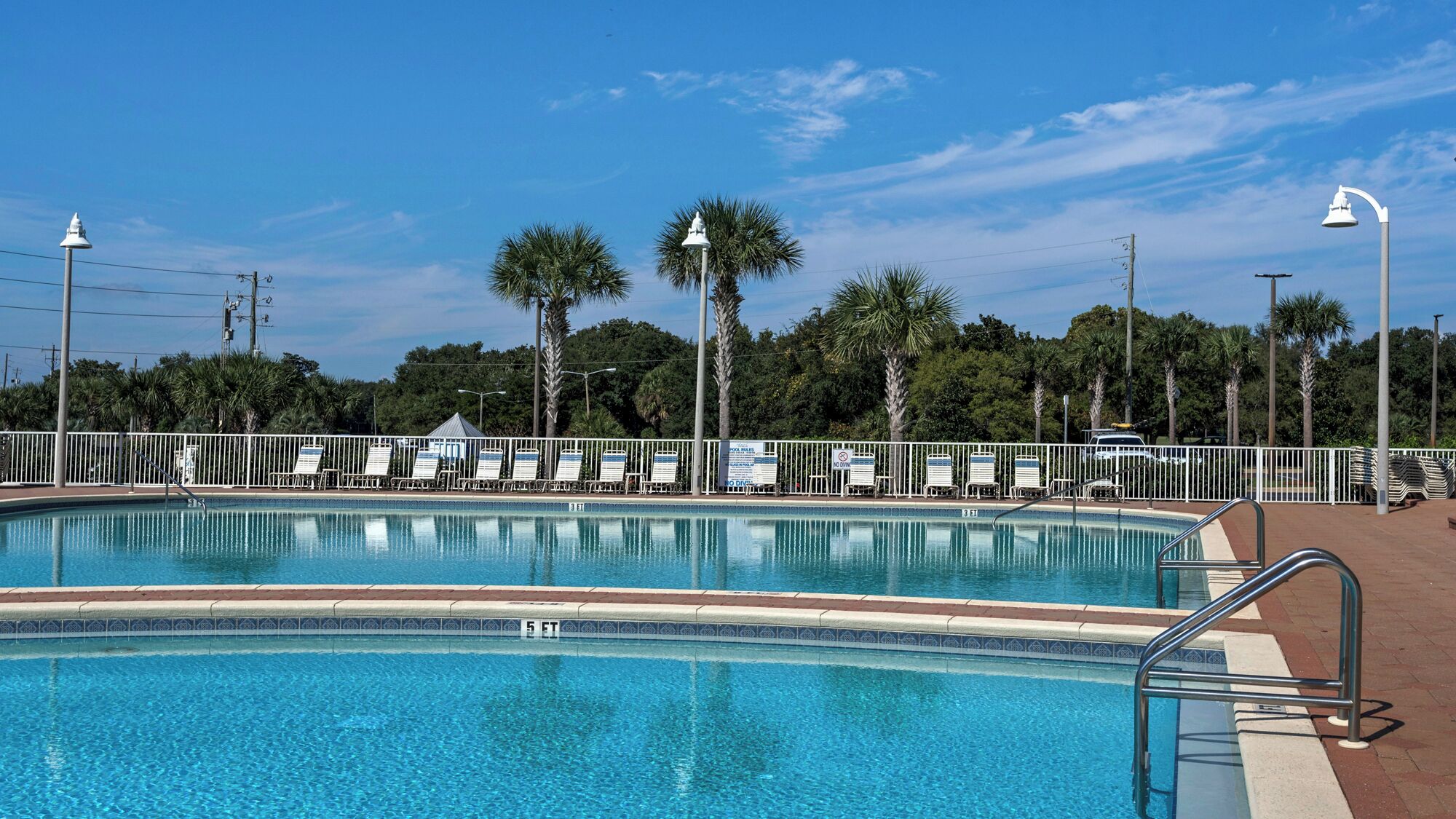 A swimming pool with a tiled edge, surrounded by a white fence, and palm trees in the background.