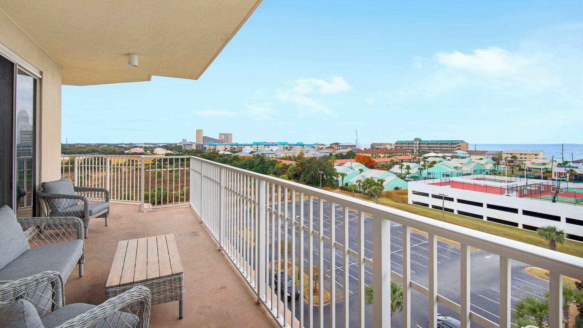A balcony with wicker furniture, a view of a parking lot, and buildings in the distance.