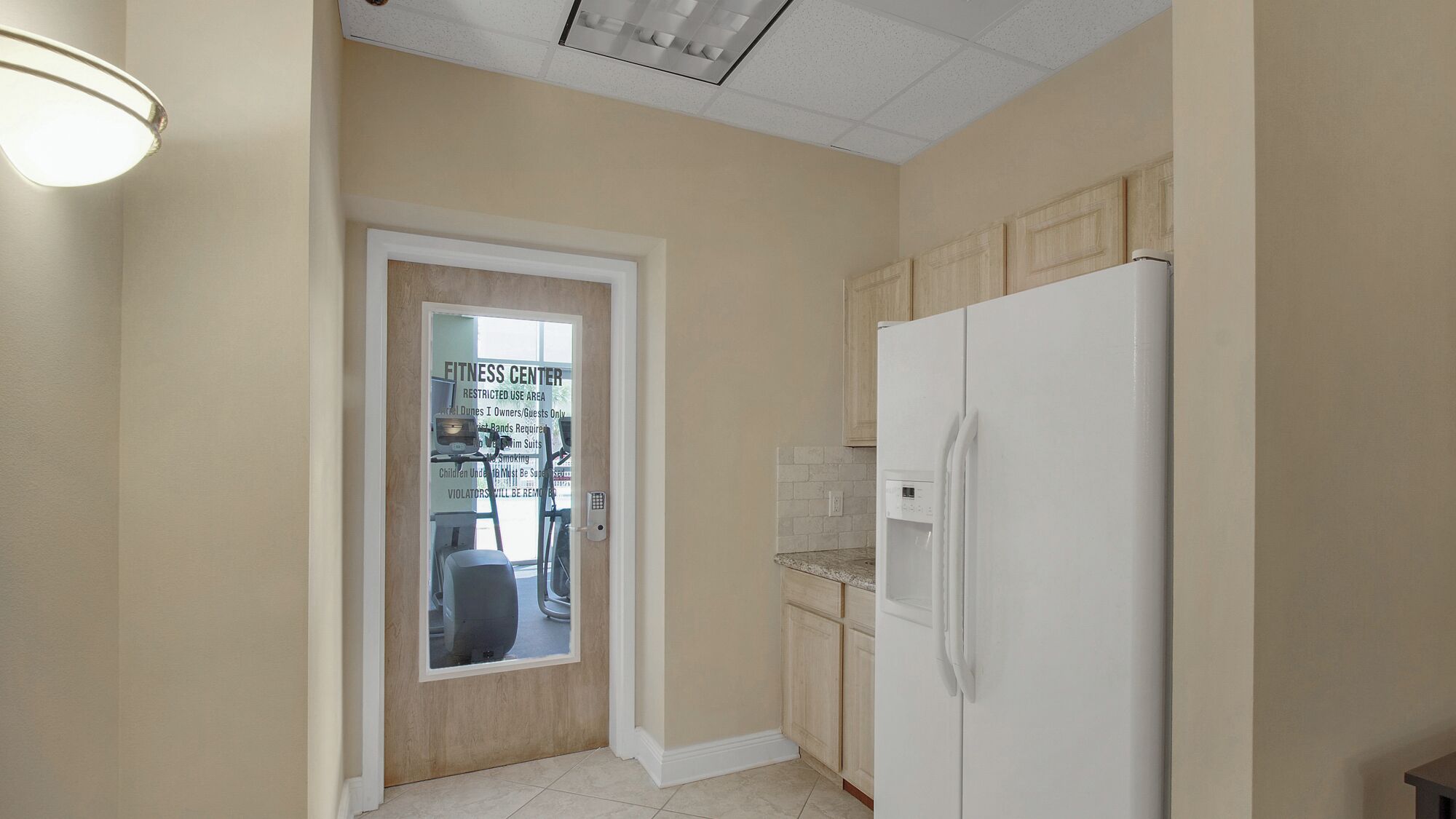 Une cuisine avec un réfrigérateur blanc, des armoires en bois et une porte donnant accès à une salle de sport.