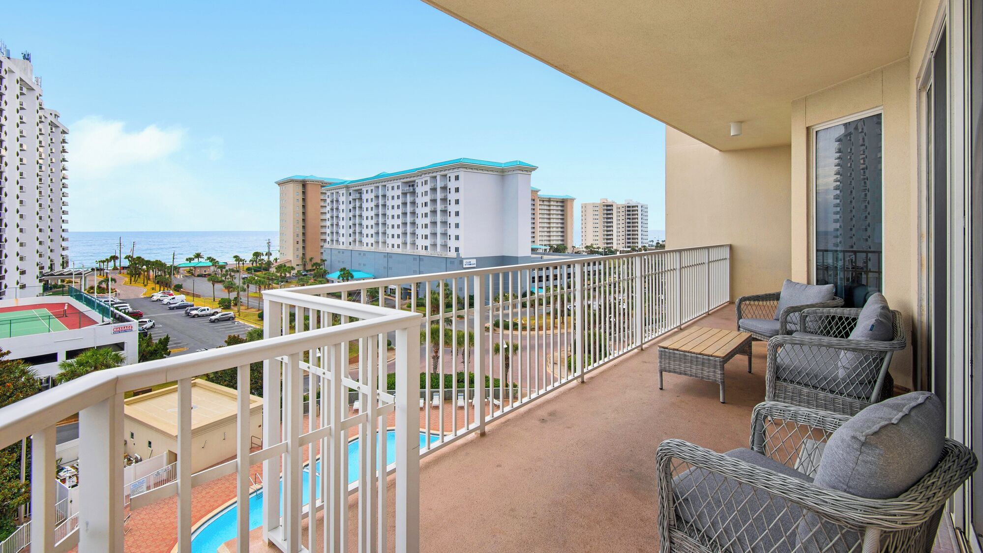 A balcony with wicker furniture, a pool, and a view of the ocean and high-rise buildings.