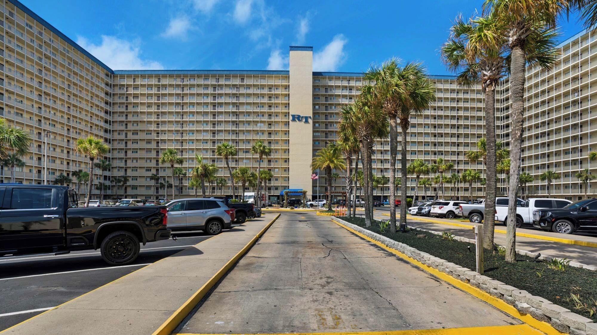A large hotel with multiple floors and a central tower. There are several parked vehicles in the foreground, and palm trees line the parking area.