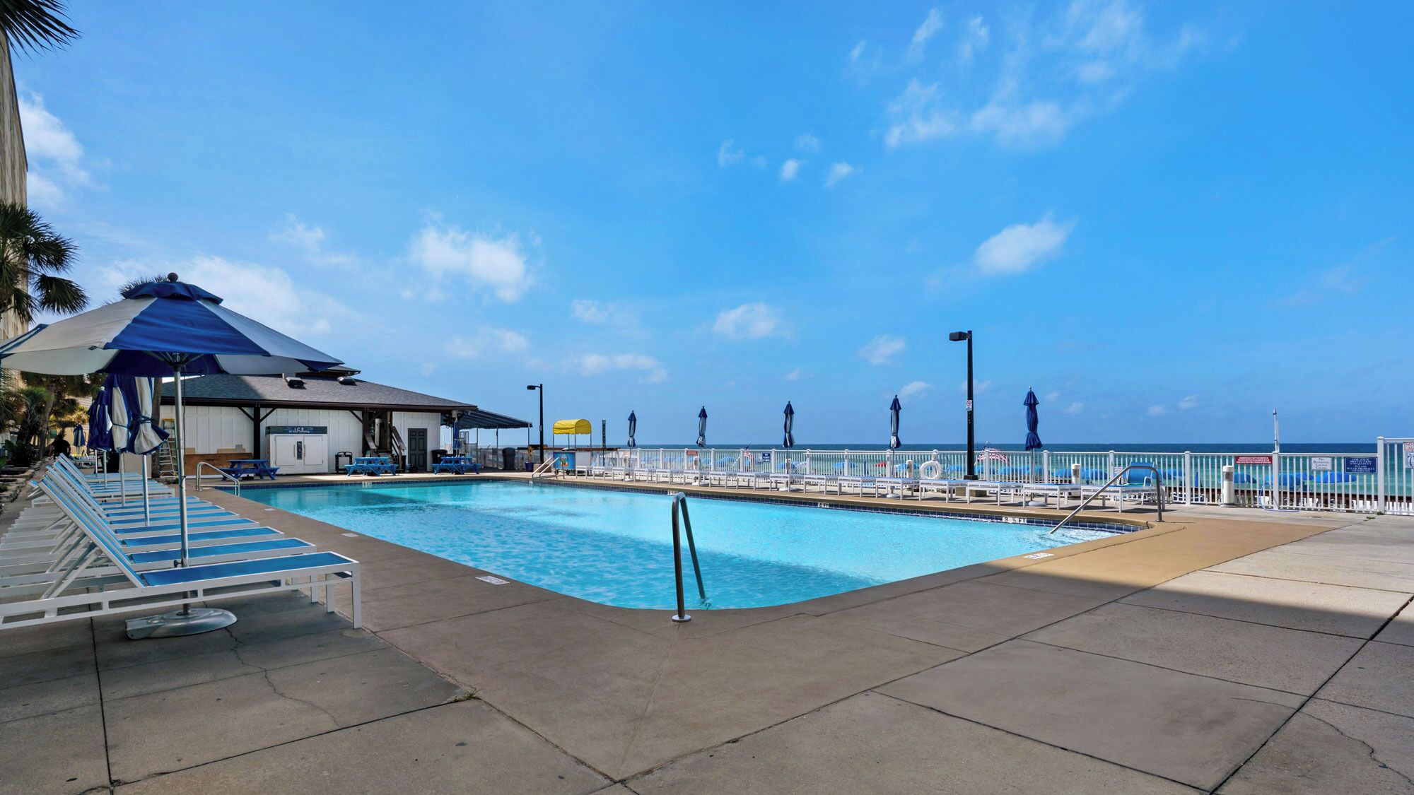 A swimming pool with lounge chairs and umbrellas, a covered area, and a view of the ocean.