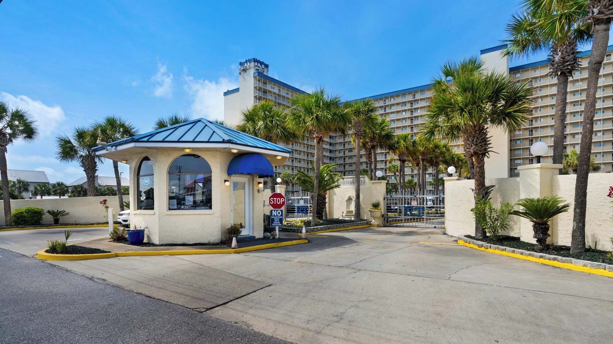 A hotel entrance with a covered waiting area, palm trees, and a gated parking area.