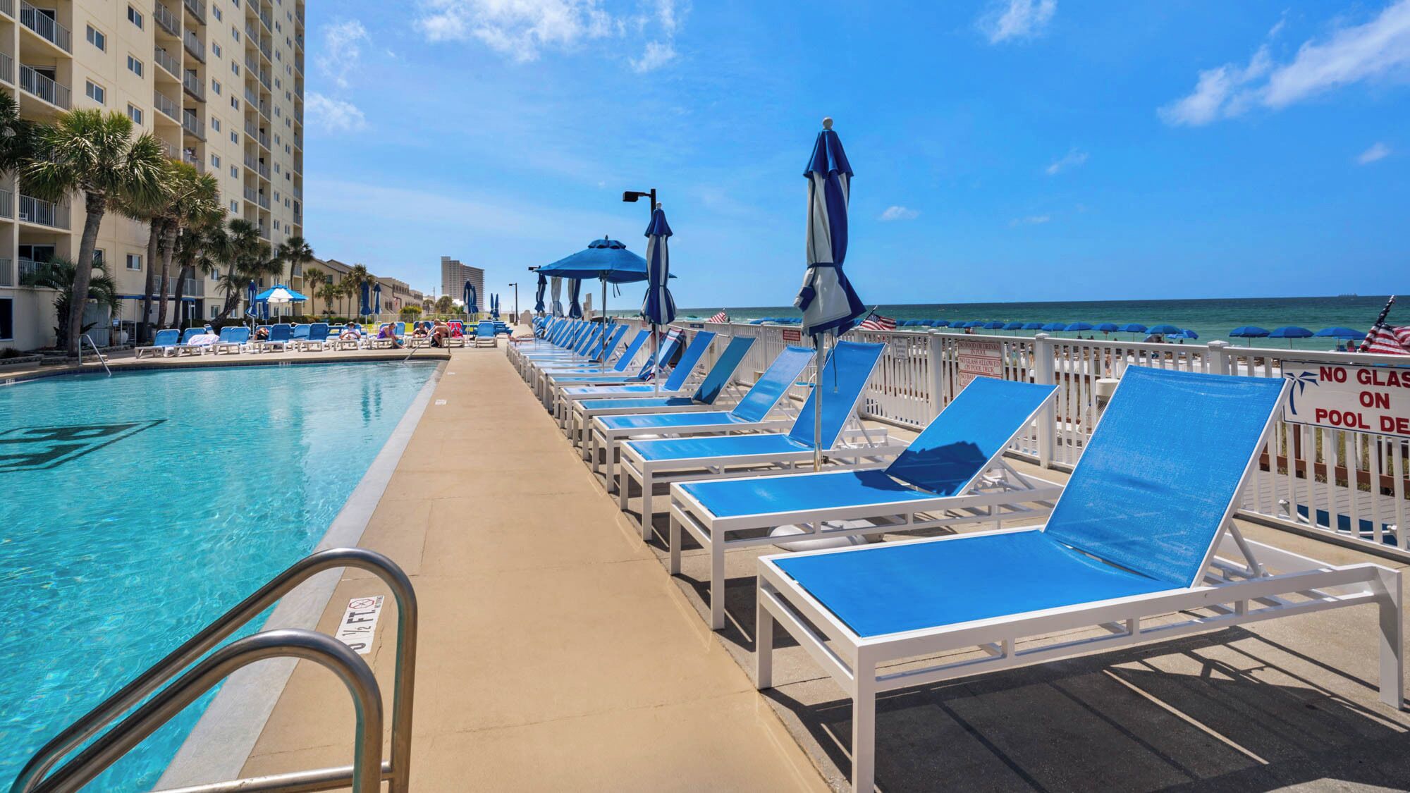 A poolside area with lounge chairs and umbrellas, a clear view of the ocean, and a building with balconies.