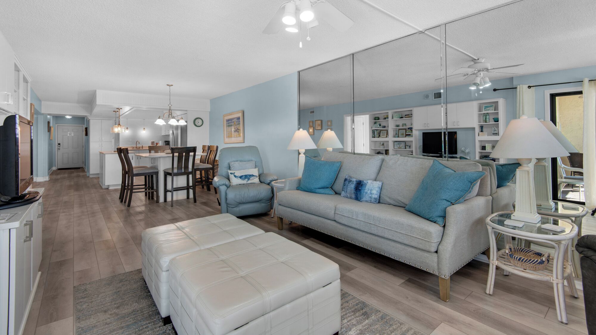 A modern living room with a grey sofa, a white ottoman, and a dining area with a kitchen in the background.