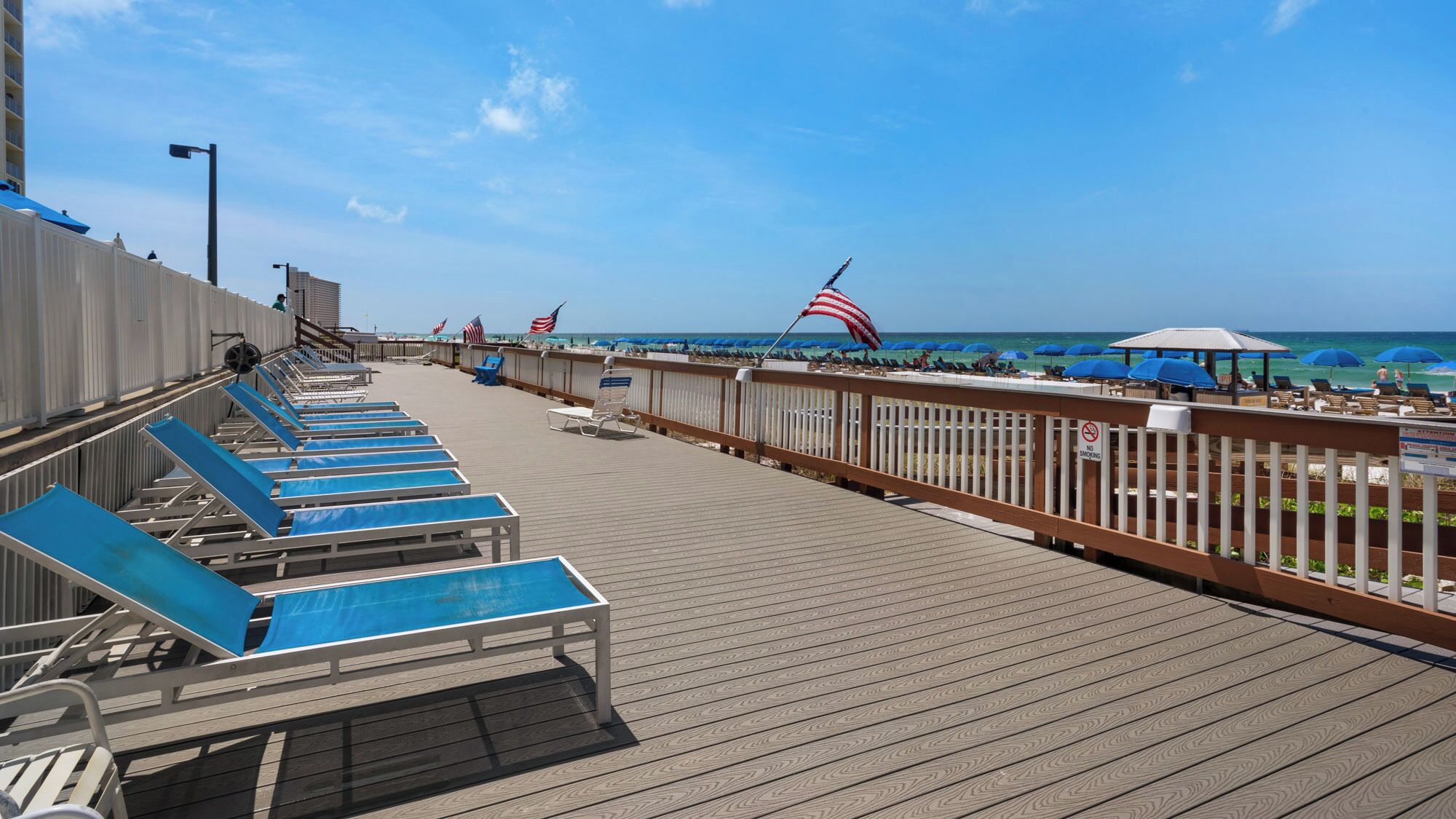 Eine Holzterrasse mit blauen Liegestühlen, einem Geländer und Blick auf den Strand mit Sonnenschirmen und amerikanischen Flaggen.