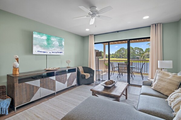 A modern living room with a large TV, a sofa, a coffee table, and a balcony with outdoor seating.