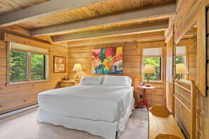 Un dormitorio de cabaña de madera con una cama, dos lámparas de noche, un cuadro abstracto colorido y un ventanal con vistas a árboles.