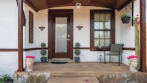 Une entrée de maison avec une porte en bois, deux plantes en pot, un banc et deux sculptures de bustes.
