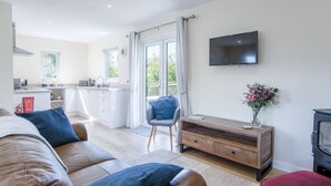 Un salon moderne avec un canapé en cuir marron, un meuble TV en bois et un pouf rouge. On aperçoit à l’arrière-plan une cuisine avec des meubles blancs et un coin repas avec une chaise bleue.