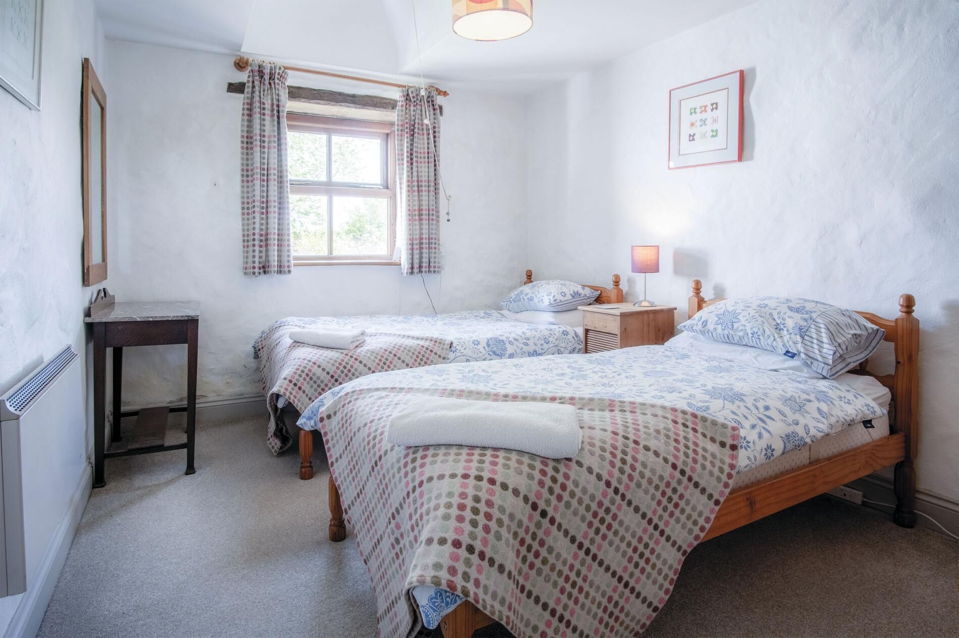 A bedroom with two single beds, a bedside table with a lamp, a window with curtains, and a framed picture on the wall.