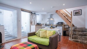 Un salon chaleureux avec un canapé et une couverture colorée, un coin cuisine doté d’appareils modernes et un escalier en pierre.