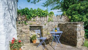 Un mur de pierres, une petite table et des chaises, une bouteille de champagne et un pot de fleurs avec des fleurs rouges.