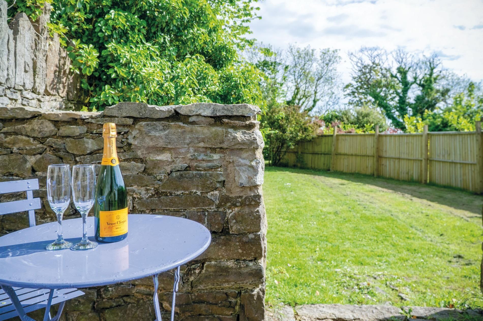 Un muro de piedra con una botella de champán Veuve Clicquot y dos copas sobre una mesa de metal.