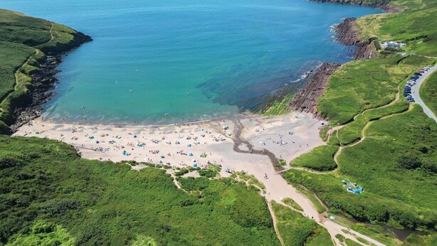 Vue aérienne d’une plage côtière, avec un rivage sablonneux, des collines verdoyantes et une mer bleue et limpide.