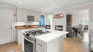A modern kitchen with white cabinetry, a central island with a stove, and a dining area with a brick accent wall. - Dog-Friendly West End Home + Parking - Sleeps 6 (Provincetown)