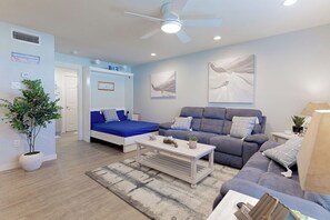 A modern living room with a blue sofa, grey sofa, and a white coffee table. There is a ceiling fan, a plant, and artwork on the walls.