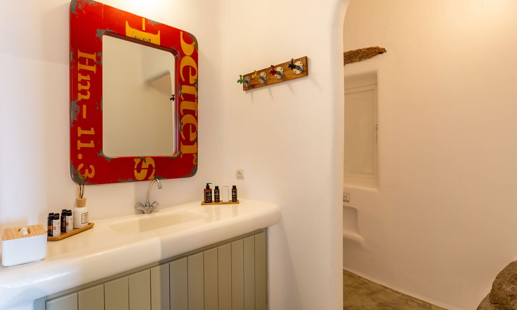 Ein Badezimmer mit weißem Waschtisch, einem roten Spiegel mit dem Schriftzug 'Hotel' und einem Holzregal mit Toilettenartikeln.
