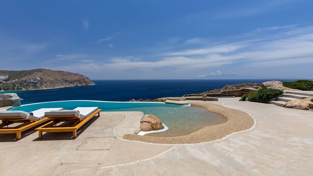 Una piscina con sillas de descanso, una playa de guijarros y vistas al mar y a las montañas.