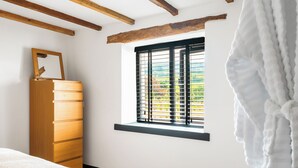 Une chambre avec une commode en bois, une fenêtre avec des stores et un peignoir blanc accroché au mur.