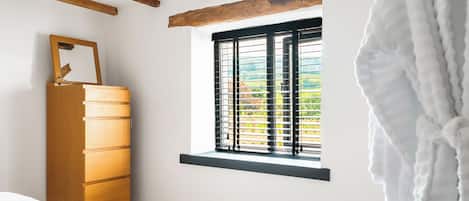 A bedroom with a wooden dresser, a window with blinds, and a white bathrobe hanging on the wall.