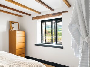 Une chambre avec une commode en bois, une fenêtre avec des stores et un peignoir blanc accroché au mur.