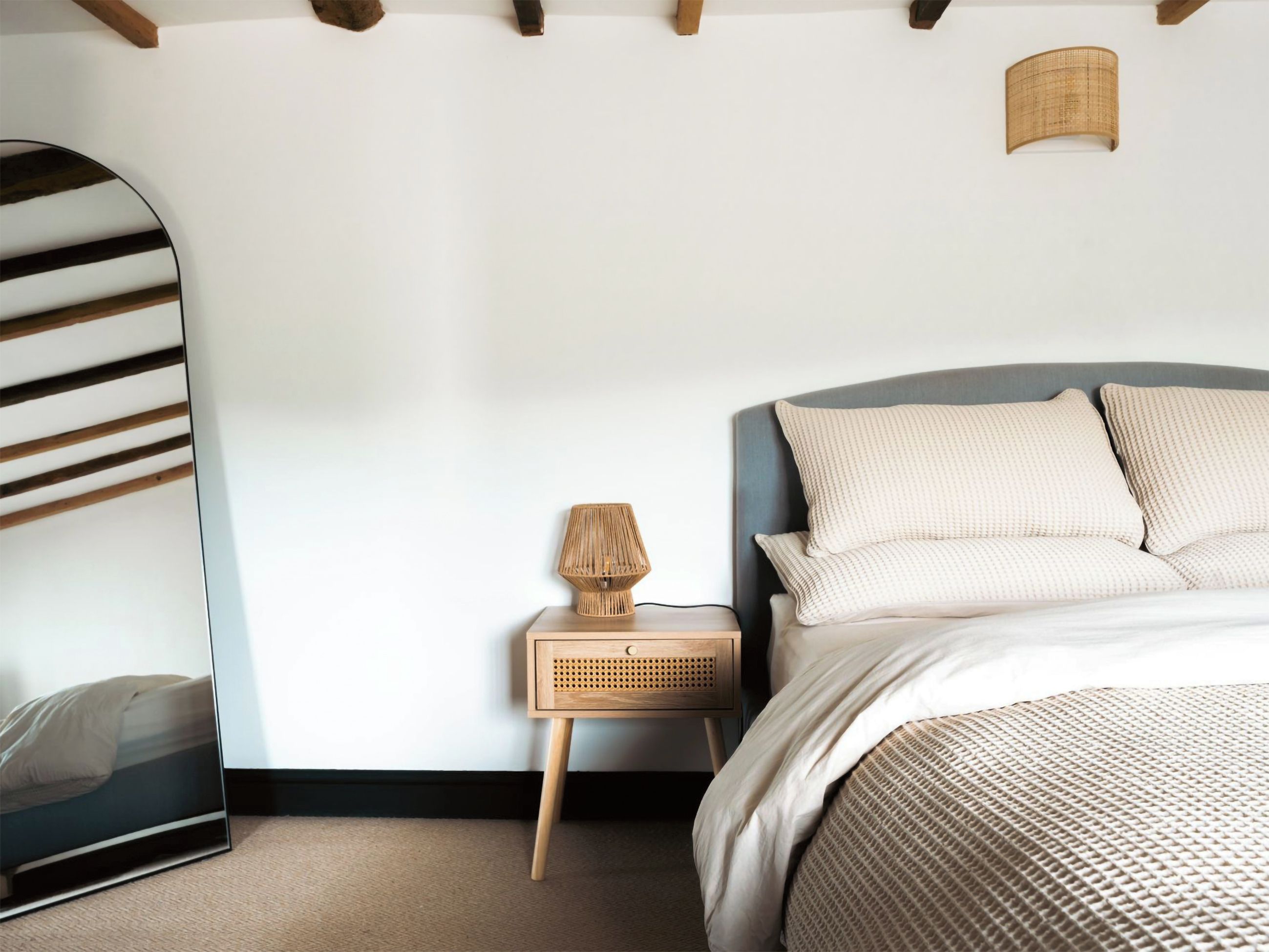 Un dormitorio con cama, mesita de noche, lámpara, espejo y una viga de techo de madera.