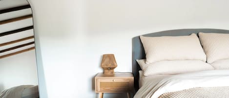 A bedroom with a bed, a nightstand, a lamp, a mirror, and a wooden ceiling beam.