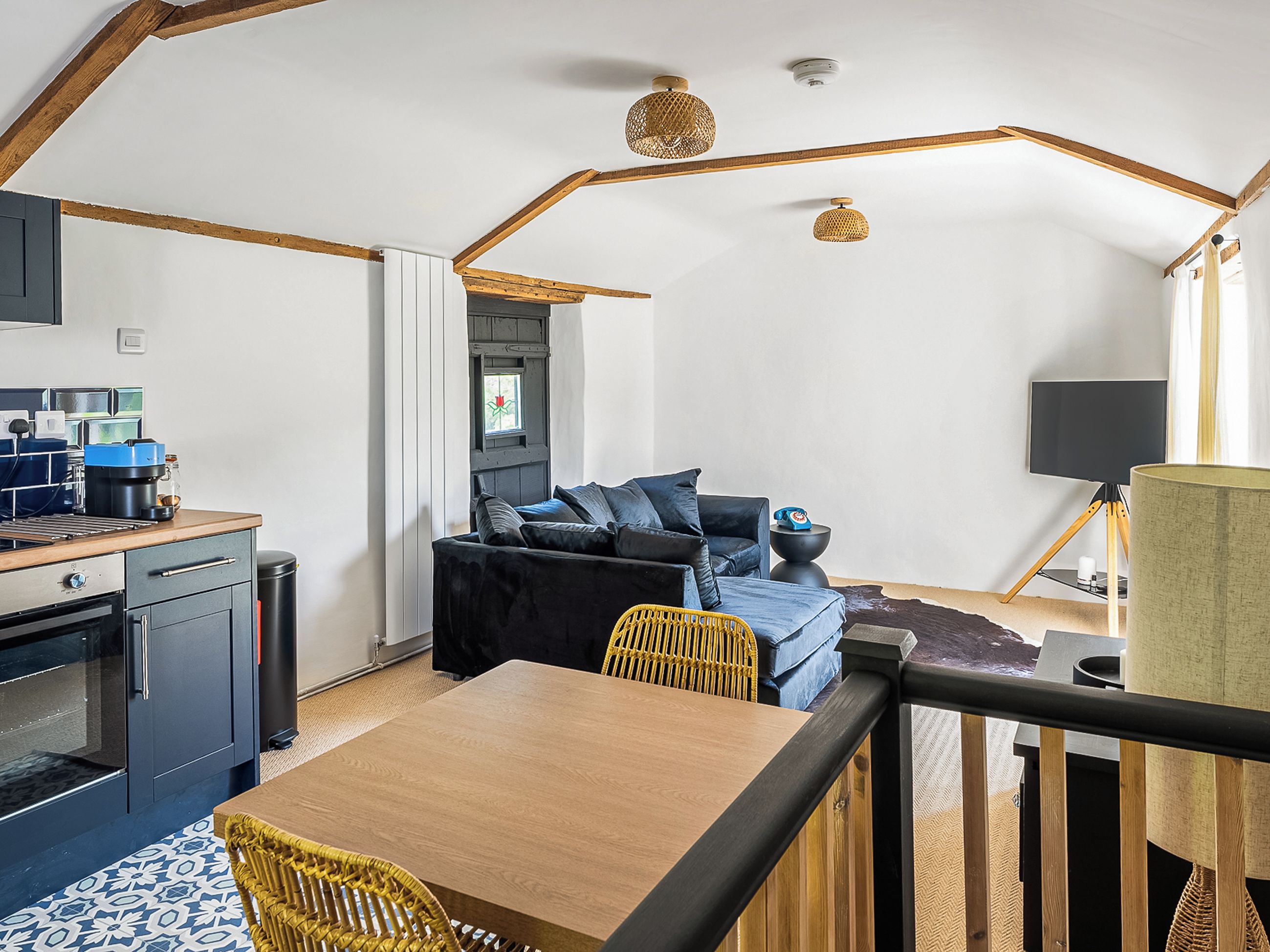 A compact kitchen with modern appliances, a dining area with a wooden table and wicker chairs, a black sofa with blue cushions, and a large window with a view.