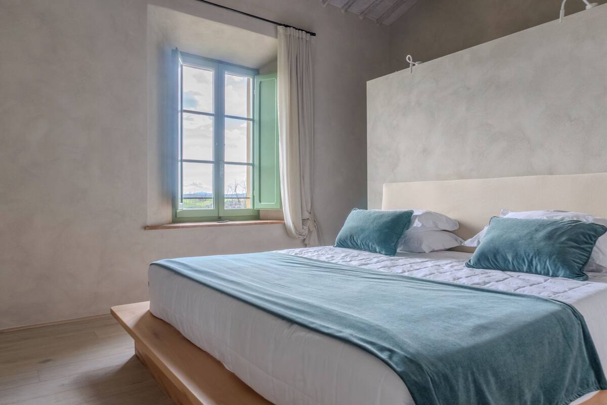 Un dormitorio con cama, ventana con cortinas, cabecero y vistas al exterior.
