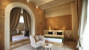 Ein Schlafzimmer mit Bett, einer Couch, einem kleinen Tisch und einem durch einen Bogenportal sichtbaren Badezimmer.