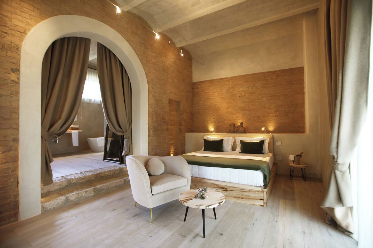 Un dormitorio con cama, sofá, una mesita y un baño visible a través de un arco.