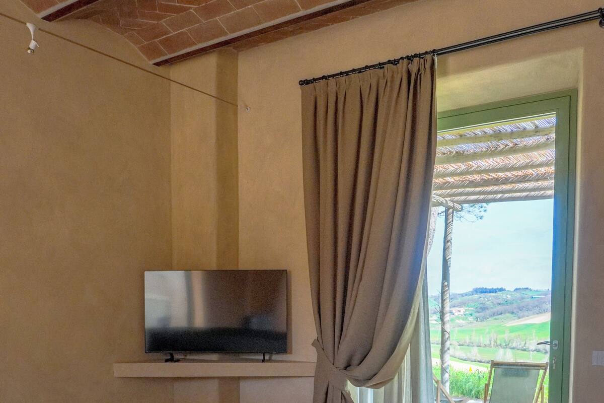 Un dormitorio con cama, televisión en un estante, cortina y vistas al exterior.