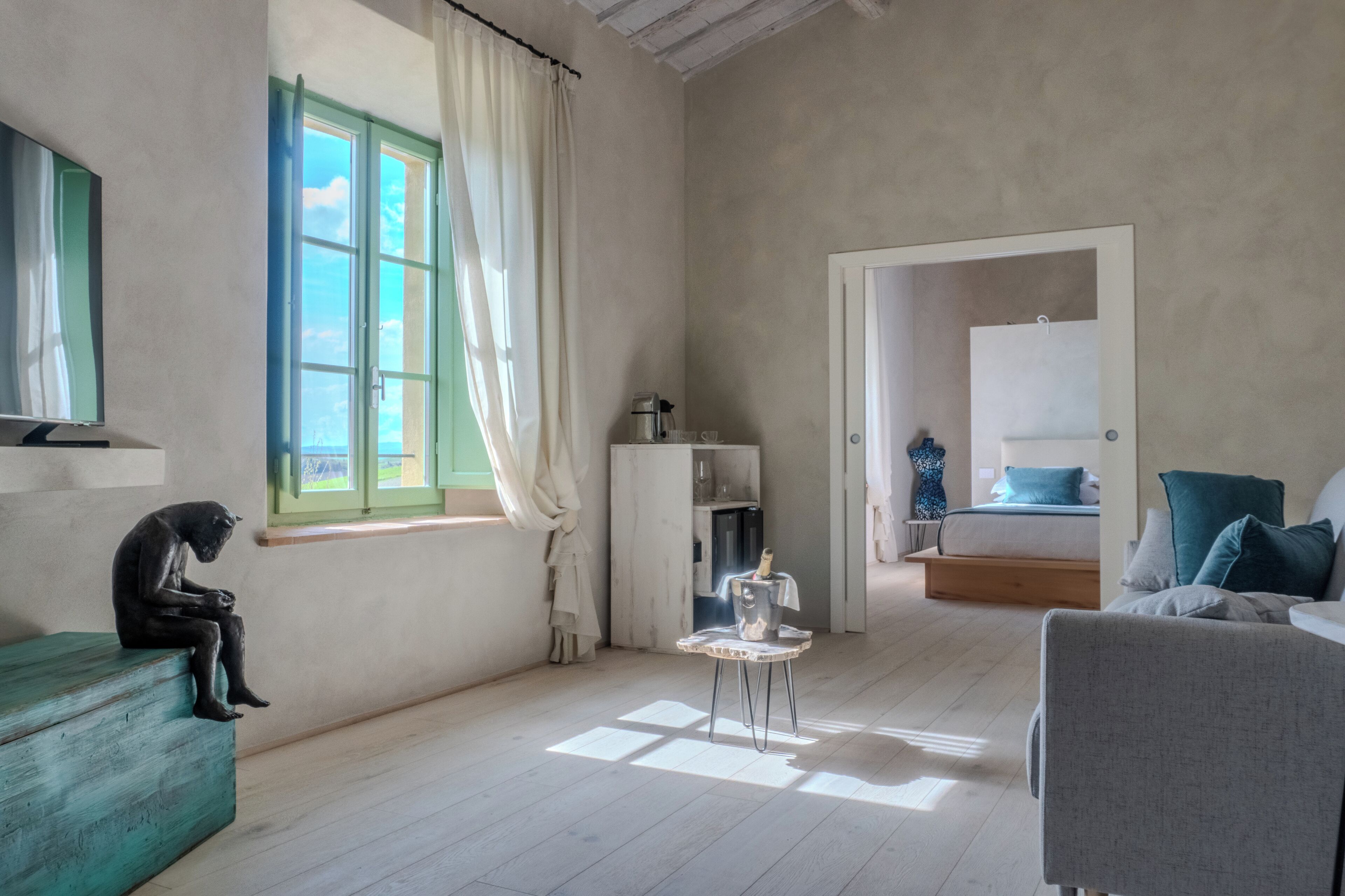 A room with a wooden floor, a green sofa, a small table with a coffee pot, a bed with blue pillows, and a window with curtains.