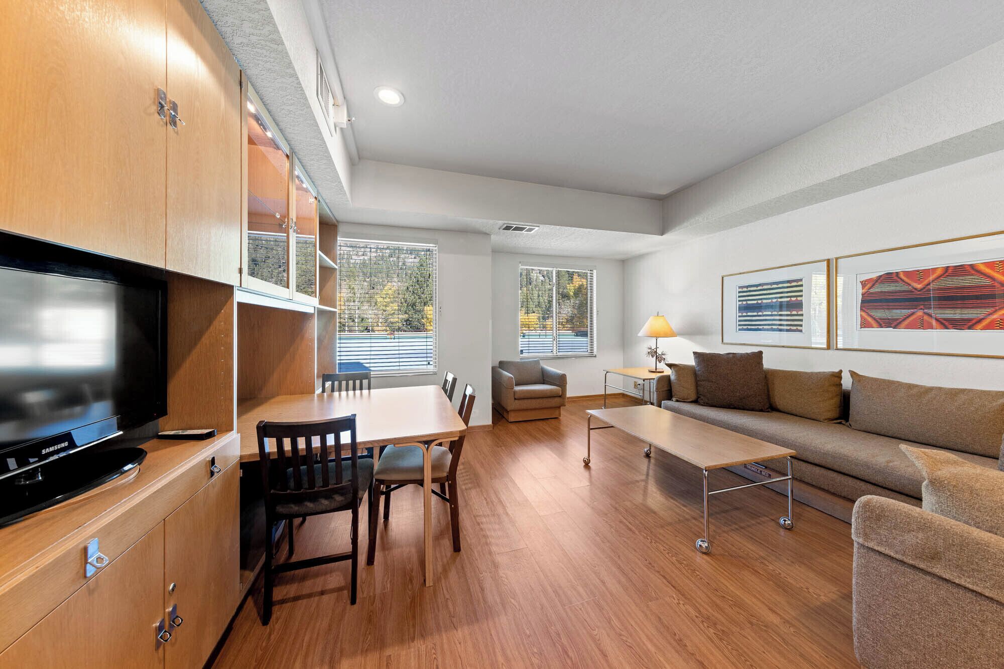 A modern living room with a flat-screen TV, wooden cabinets, a dining table with chairs, a sofa, and two windows with blinds.