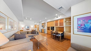 A modern hotel room with a sofa, a small table, a TV, and a kitchen area with wooden cabinets and a dining table. - 229 | Studio w/ AC unit - Stroll to the Village (Olympic Valley)