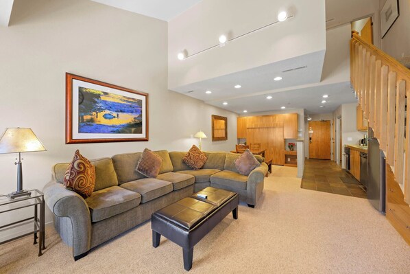 A spacious living room with a sectional sofa, a coffee table, and a dining area with wooden cabinets. - Fresh Tracks Loft | Top-Floor Loft, Hot Tubs, 348 (Olympic Valley)