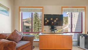 A cozy living room with a large flat-screen TV, a wicker sofa, and a wooden cabinet. - 348 | Refreshed Loft - Full Fridge + Pool Vibes (Olympic Valley)
