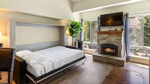 Ein Schlafzimmer mit Bett, Kamin aus Stein, Wand-Fernseher und großem Fenster mit Vorhängen.