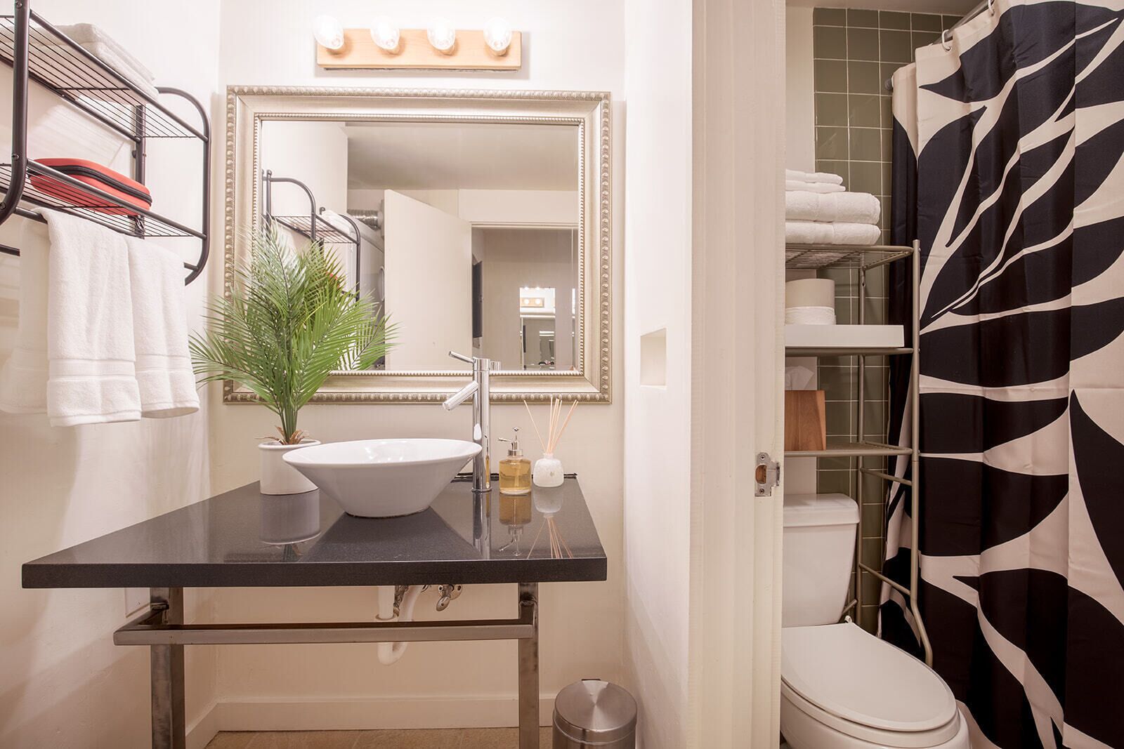 A bathroom with a black countertop, white sink, and a mirror above it. There is a plant on the countertop, a towel rack on the wall, and a shower curtain with a black and white pattern.