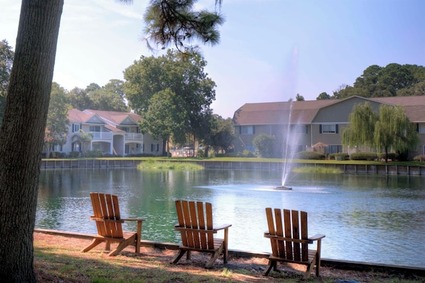 A tranquil lakeside setting with three wooden Adirondack chairs facing a pond, a fountain, and a row of houses in the background. - Renovated 2br/2ba Ocean Walk. Walking distance to pier/Village area (Saint Simons Island)