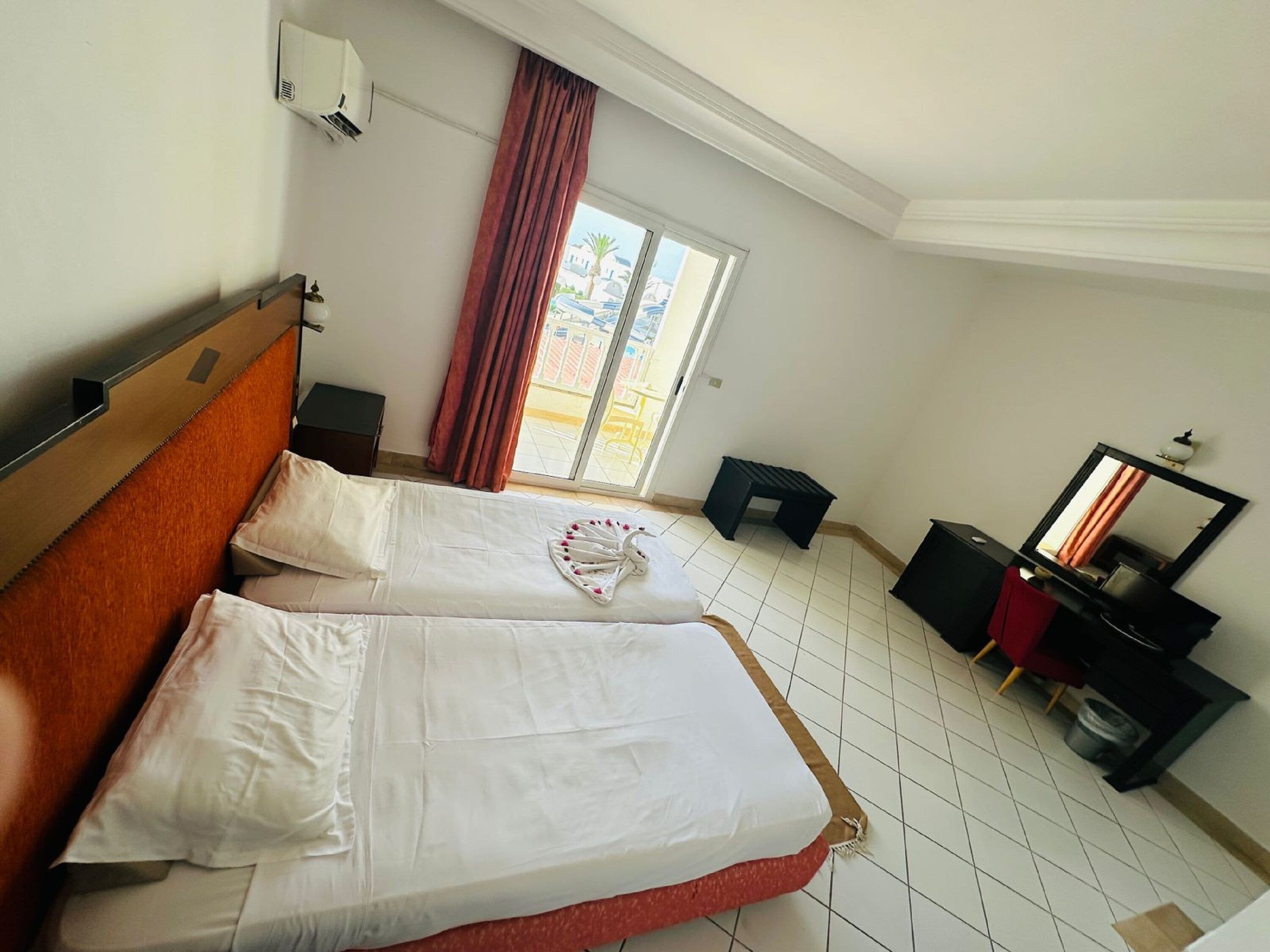 A hotel room with two beds, a large window with curtains, a small table, a mirror, and a TV.